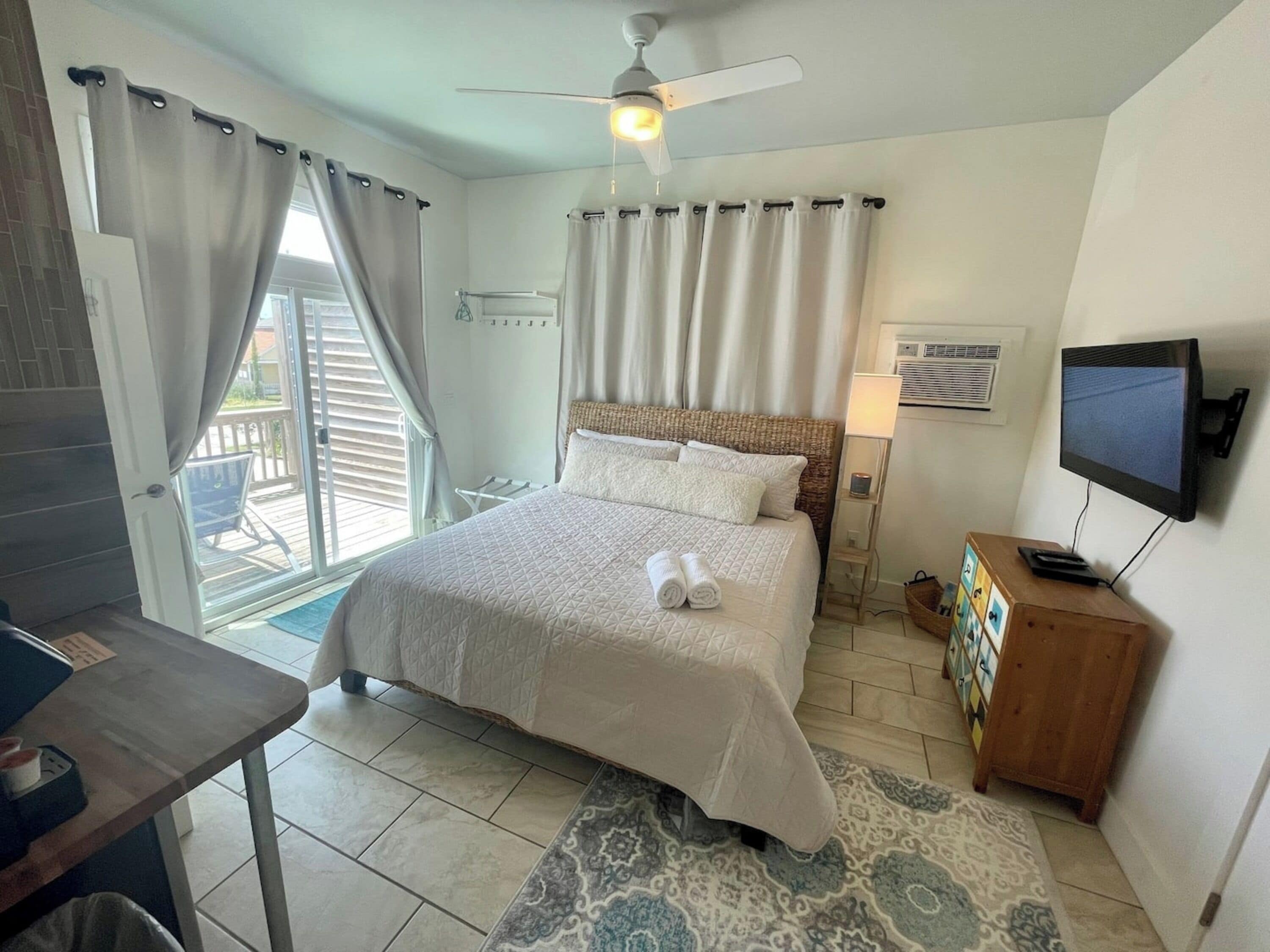 A hotel room with a bed, a TV, a ceiling fan, a desk, and a window with curtains.