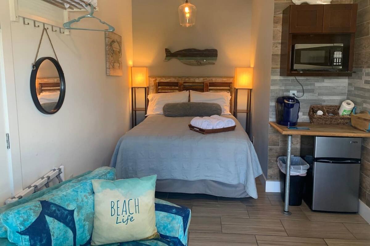 Une chambre d’hôtel compacte comprenant un lit, des lampes de chevet, une petite kitchenette, un plancher en bois et une décoration murale représentant des poissons.
