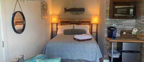 A compact hotel room with a bed, bedside lamps, a small kitchenette, a wooden floor, and a wall-mounted fish decoration.