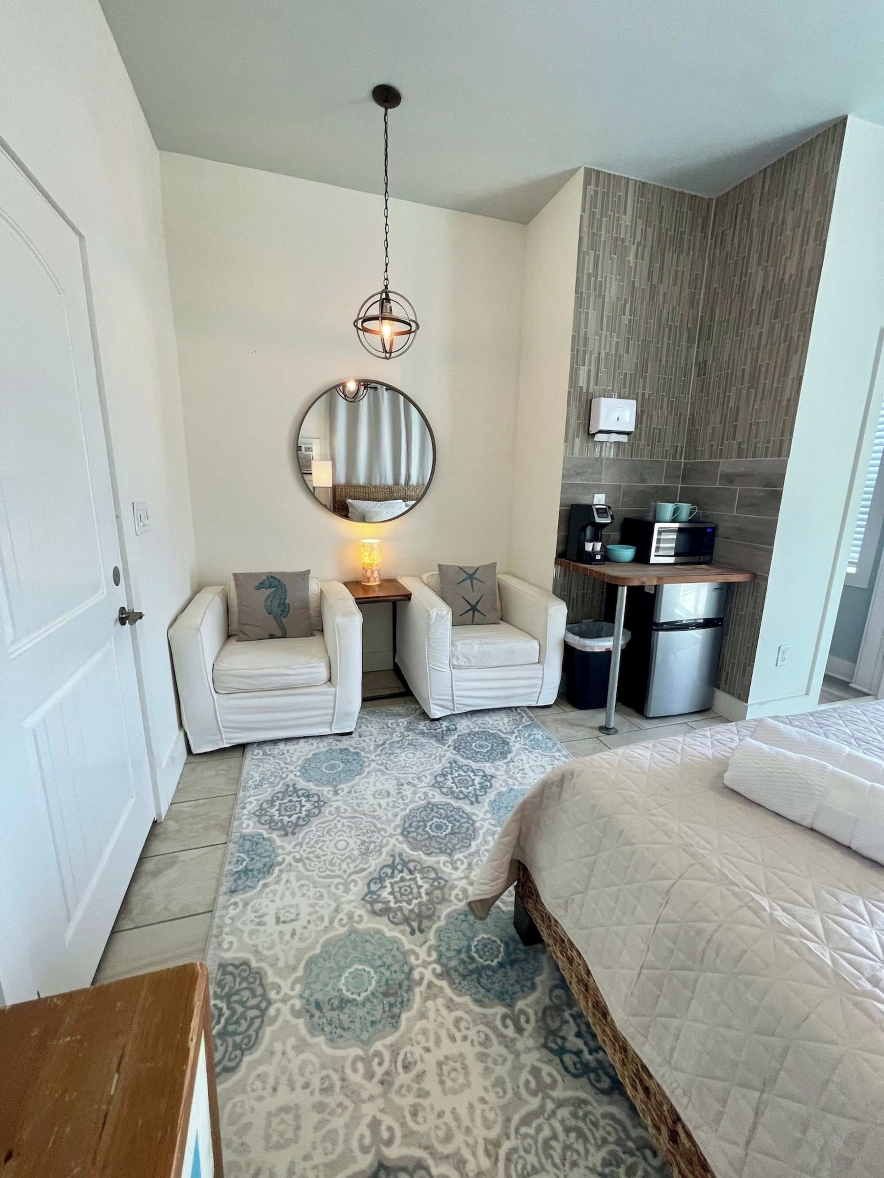 A hotel room with a bed, two armchairs, a small table with a lamp, a mirror, and a kitchenette with a microwave and refrigerator.