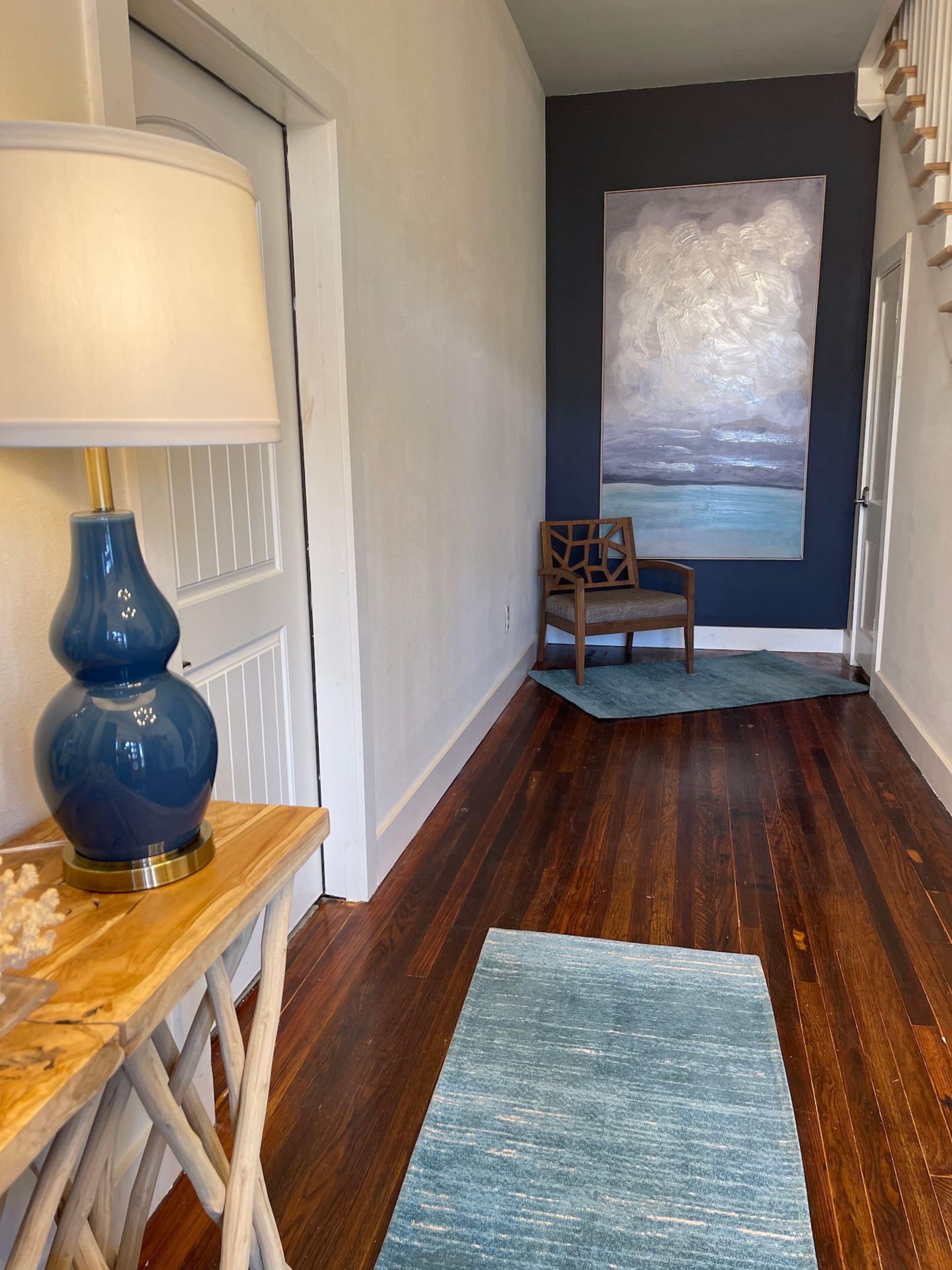 Een hal met een houten vloer, een blauw vloerkleed, een stoel, een lamp en een schilderij aan de muur.