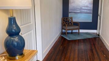 A hallway with a wooden floor, a blue rug, a chair, a lamp, and a painting on the wall.