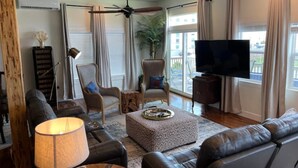 Un salon comprenant un canapé, un fauteuil, un pouf, un téléviseur, un ventilateur de plafond et une plante.