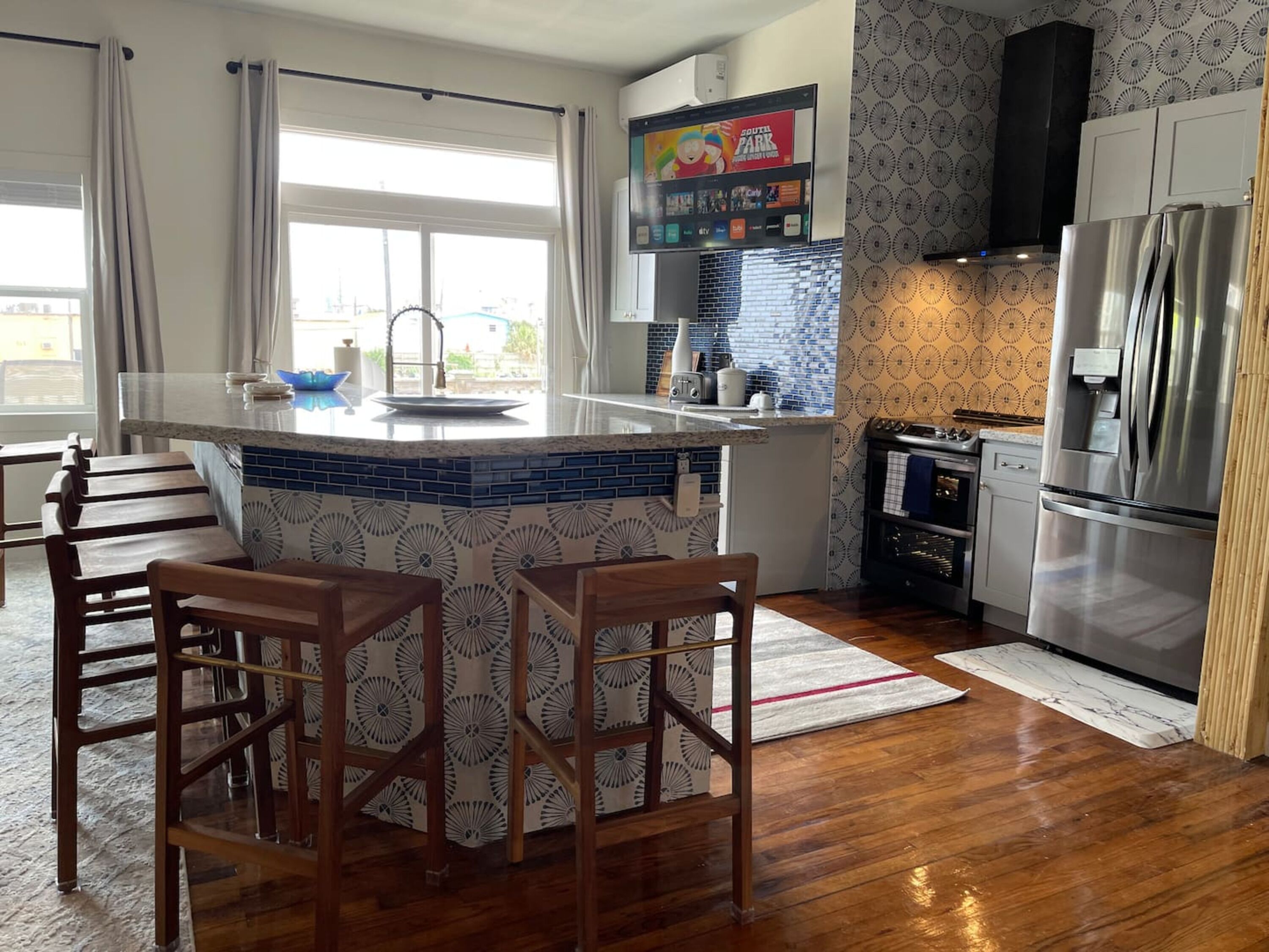Una cocina con una isla central, taburetes de bar, un refrigerador y un televisor montado sobre la isla.