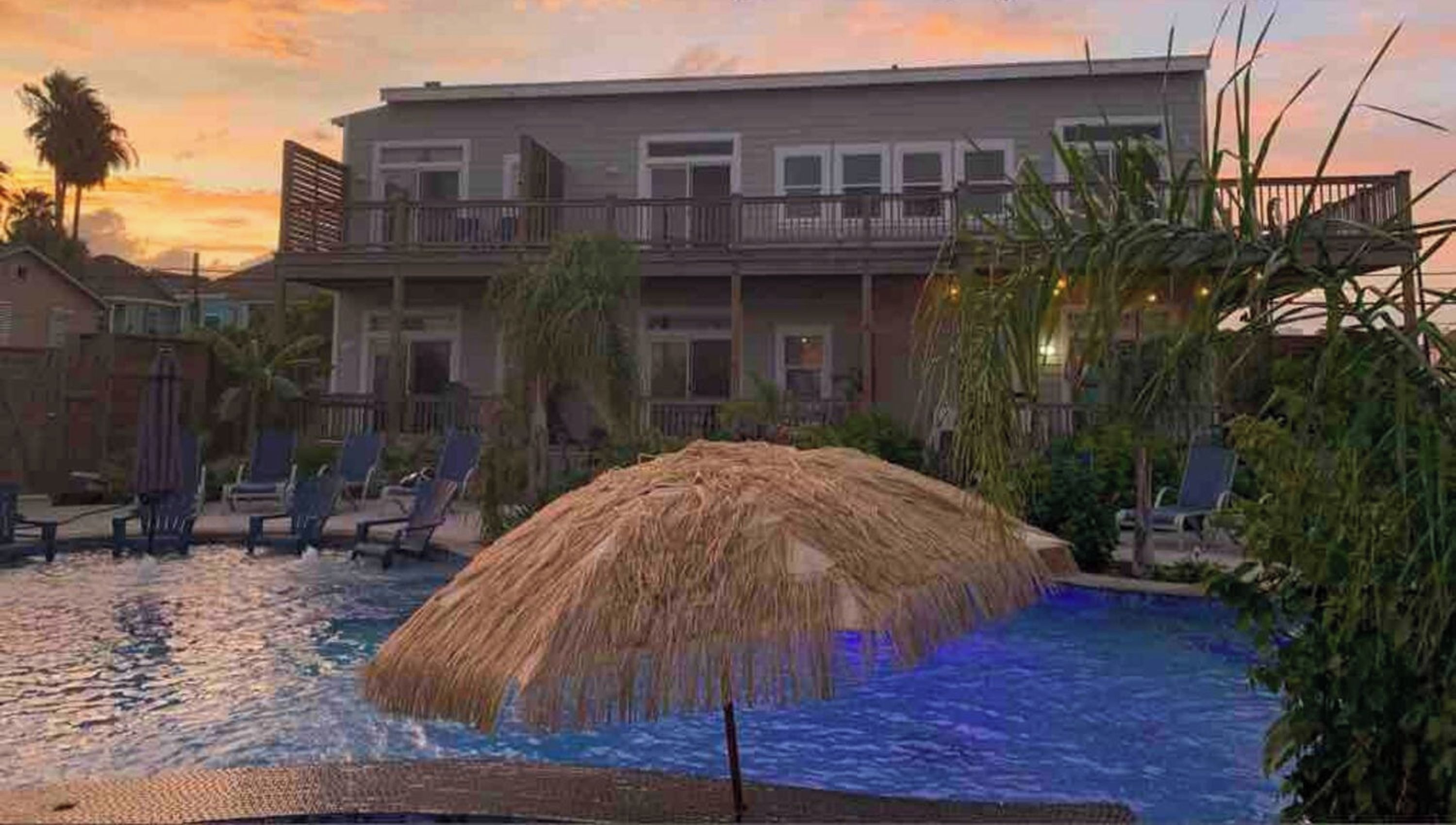 A pool with a thatched umbrella, lounge chairs, and a building with balconies in the background.