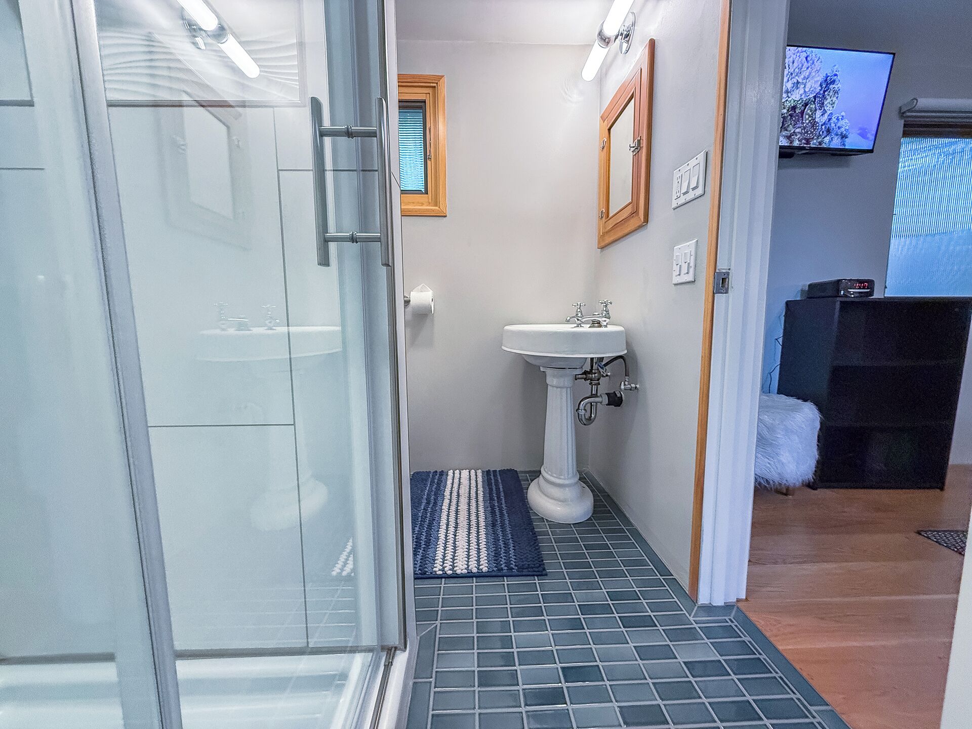 A bathroom with a glass shower enclosure, a pedestal sink, a small window, and a television on the wall.