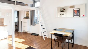 A room with a loft bed, a desk with two chairs, a ladder, and a small shelf with items. - Cozy apartment with loft bed in Dresden Neustadt - your temporary home (Dresden)
