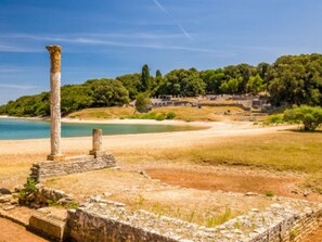 An ancient ruin with stone columns and walls, a beach, and a forested hill in the background. - Cozy Apartment With Patio and Parking (Fazana)