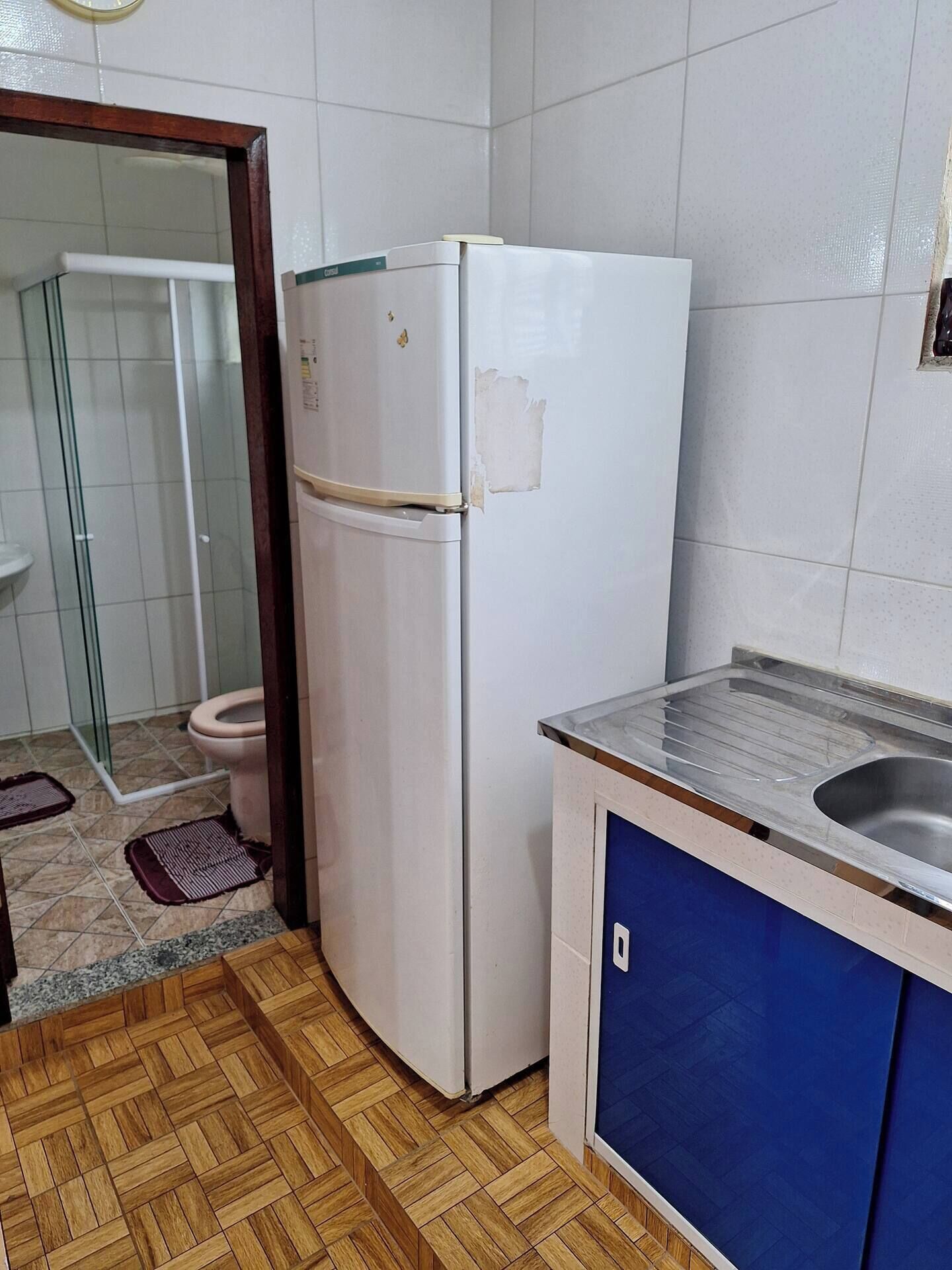 A compact kitchen with a white refrigerator, a sink with a blue cabinet, and a shower area visible through a glass door.