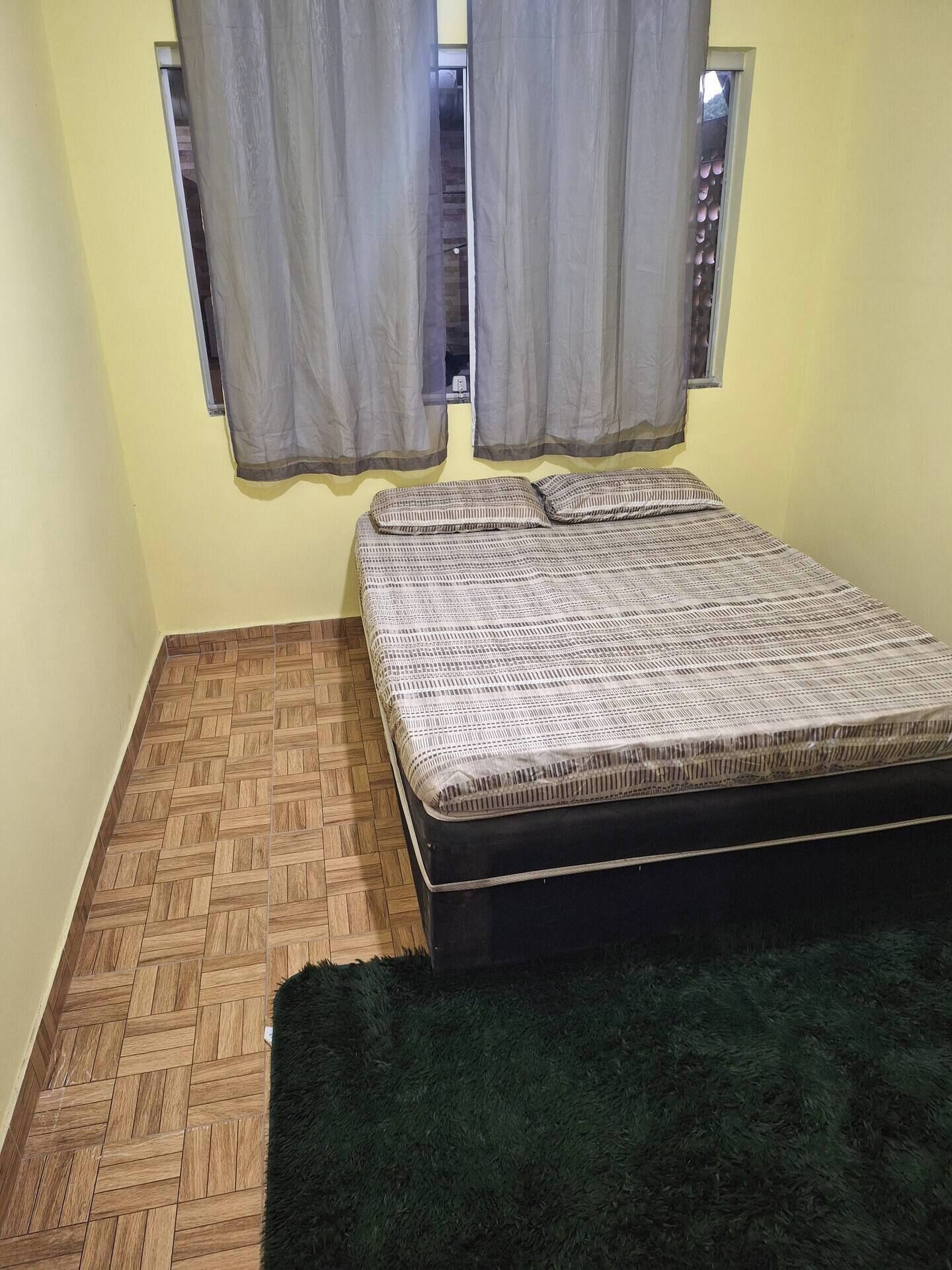 Una habitación pequeña con una cama, una ventana con cortinas, piso de madera y una alfombra verde.