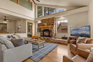A spacious living room with a stone fireplace, a large TV, and a high ceiling with skylights.