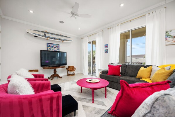 Una sala de estar moderna con un televisor de pantalla plana, ventilador de techo, un ventanal con cortinas y una decoración con una tabla de surf.
