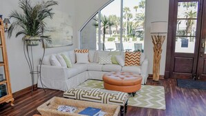 A modern living room with a white sectional sofa, a round orange ottoman, a woven coffee table, and a large window offering a view of palm trees.