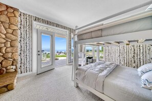 A bedroom with a bunk bed, a stone wall, a door leading to a balcony, and a view of the landscape.