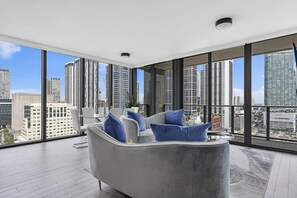 Un moderno salón con un sofá gris, cojines azules y vistas a la ciudad a través de amplias ventanas.