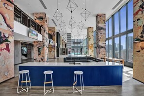 A modern hotel lobby with a central bar area, high ceilings, and large windows offering city views.