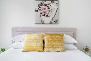 A bed with white bedding and two gold-striped pillows, a grey headboard, and a framed floral artwork above.