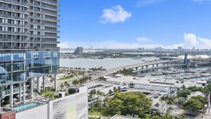 A modern high-rise hotel with a glass facade, a rooftop pool, and a view of a cityscape including a bridge and marina. - X Lounge V-Panoramic Private Residence (Miami)
