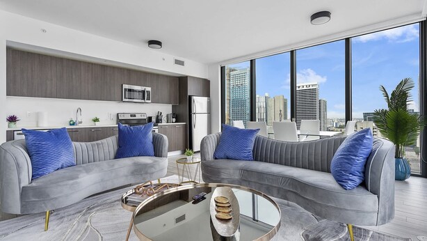 A modern living room with two grey sofas, a glass coffee table, and a city view through large windows. - X Lounge V-Panoramic Private Residence (Miami)