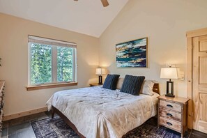 A bedroom with a large bed, two bedside lamps, a painting on the wall, and a window with a view of trees. - Twin Creek Lodge (Breckenridge)