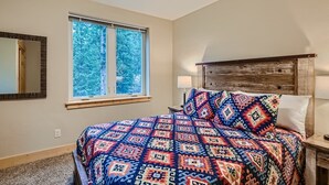 A bedroom with a large bed, a window with a view of trees, a wooden headboard, and a bedside table with a lamp.