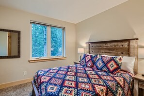 Une chambre à coucher avec un grand lit, une fenêtre donnant sur les arbres, une tête de lit en bois et une table de chevet avec une lampe.