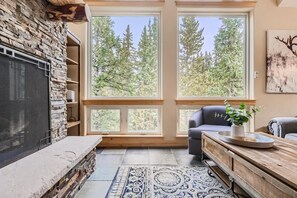 A cozy living room with a stone fireplace, large windows with a forest view, and a comfortable seating area. - Twin Creek Lodge (Breckenridge)