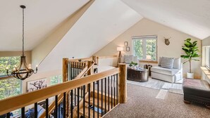 A spacious loft with a high ceiling, a wooden staircase, a cozy seating area with a sofa and armchairs, and a large window offering a view of the outdoors.