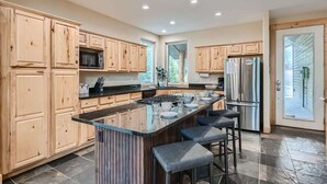 A modern kitchen with wooden cabinets, a central island with bar stools, stainless steel appliances, and a large window providing natural light.