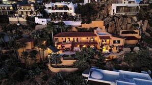 A large, multi-story building with a red-tiled roof, surrounded by palm trees and rocky terrain.