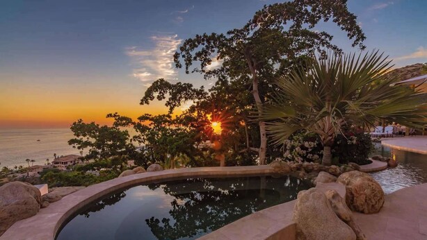 A pool with a curved edge, surrounded by rocks and palm trees, with a sunset view over the ocean.