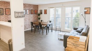 Een moderne woonkamer met een grijze bank, een houten eettafel met stoelen, een accentmuur van baksteen en een balkon met uitzicht.