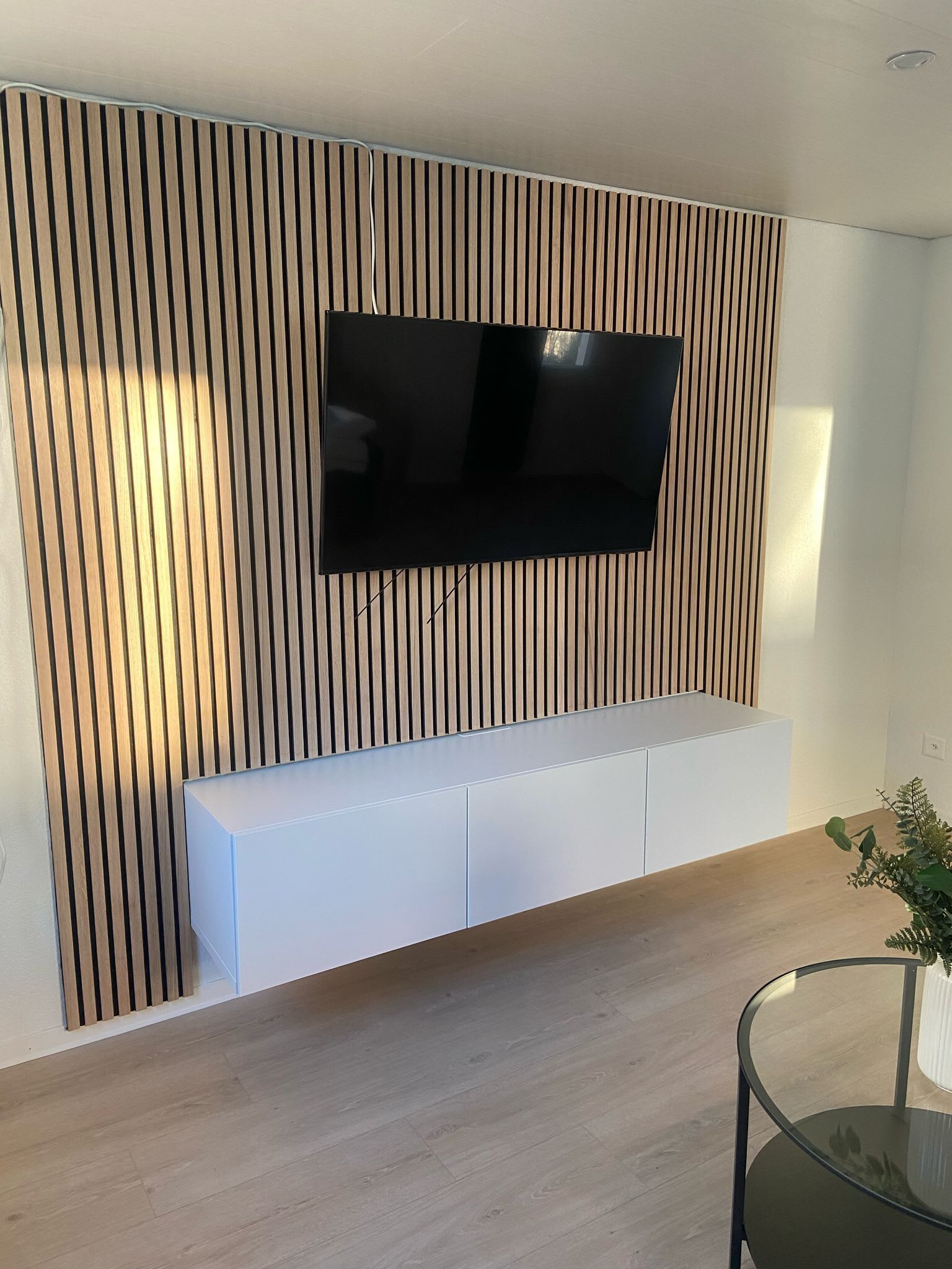 A modern living room with a flat-screen TV mounted on a wooden slat wall, a white built-in bench, and a glass coffee table.