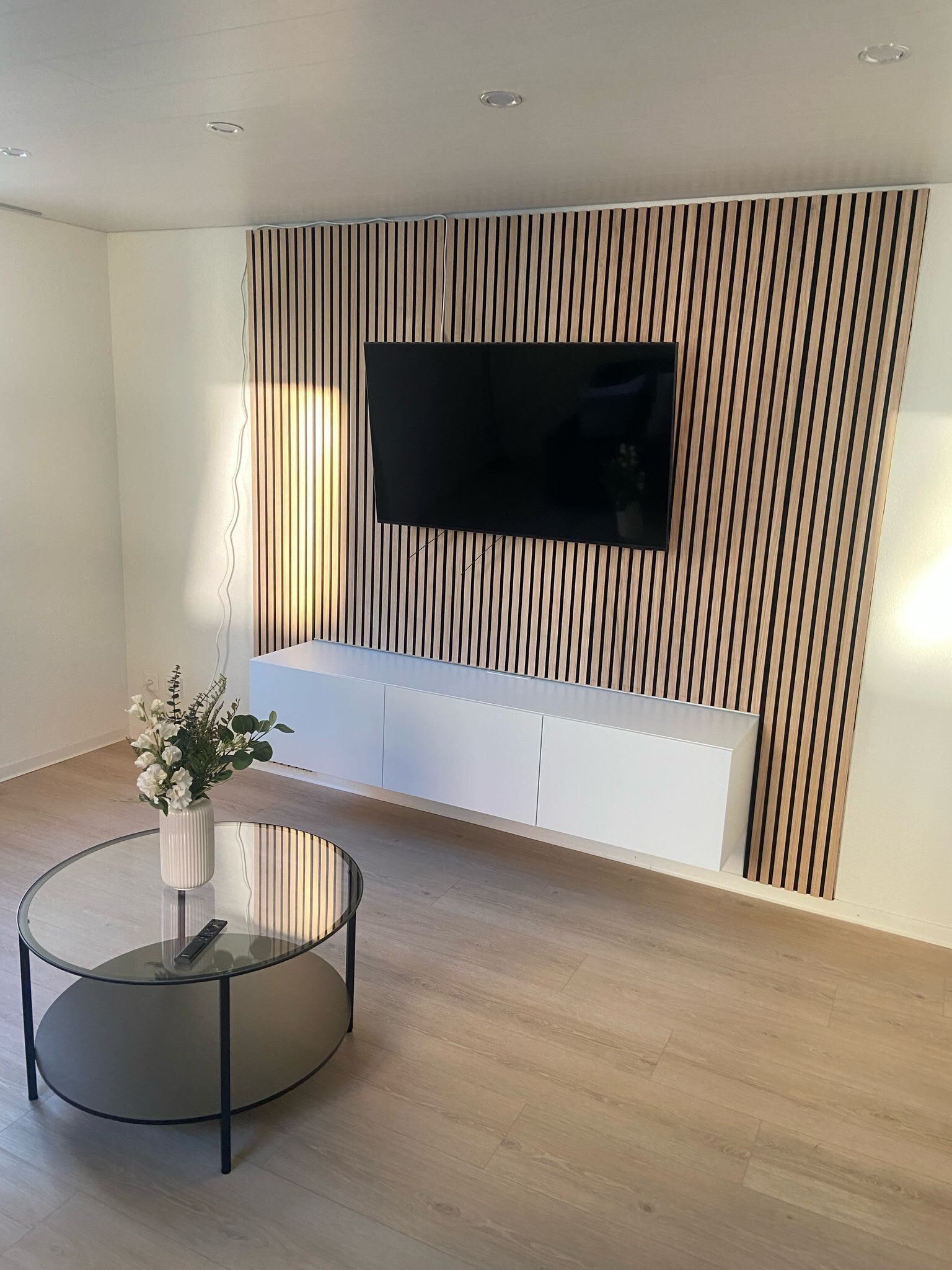 Un soggiorno moderno con una TV a schermo piatto montata su una parete rivestita in legno, un tavolino da caffè in vetro e un vaso di fiori, e un mobile TV bianco sottostante.