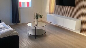 Habitación de hotel moderna con televisor de pantalla plana montado en una pared de paneles de madera, una mesa redonda de cristal con un florero, una cama con ropa blanca y una ventana con contraventanas rojas.