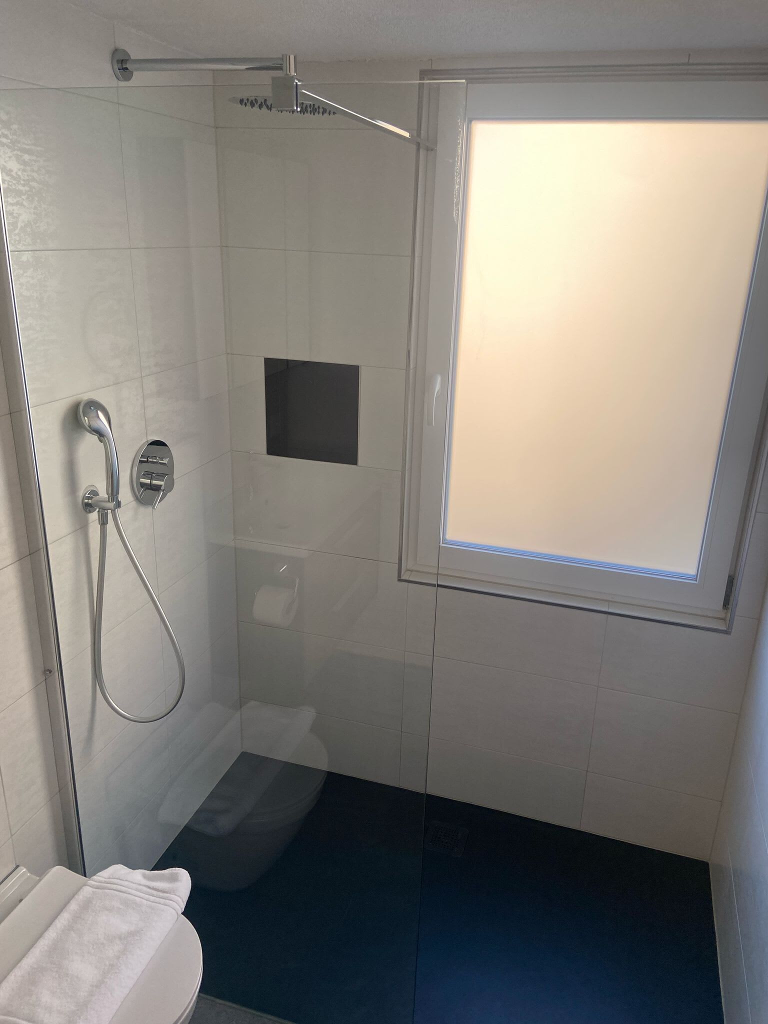 Un petit espace douche fermé, doté d’une porte en verre, d’une serviette blanche posée sur les toilettes et d’une fenêtre à vitrage dépoli.