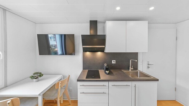 A compact kitchen with white cabinets, a dark countertop, and a built-in stove. - book2sty: Cozy Studio with kitchenette and washing machine (Herisau)
