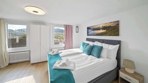 A hotel room with a bed, a window with a view, a painting on the wall, and a nightstand with a lamp.