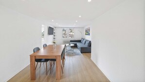 A modern living space with a wooden dining table, grey sofa, and a view of the city through a window.