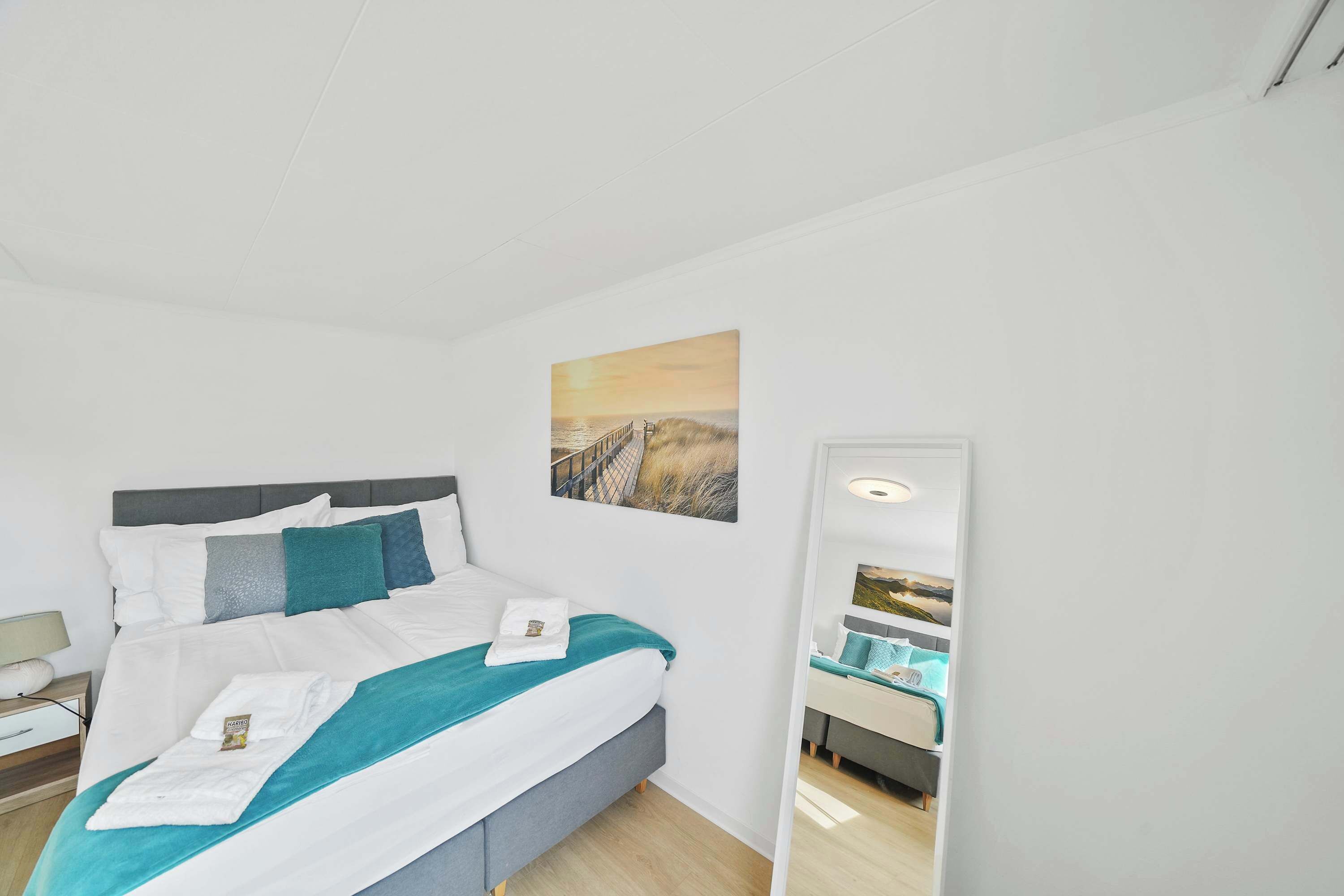 Uma cama bem arrumada com roupa de cama branca e azul-turquesa, uma mesa de cabeceira com abajur e um quadro de pôr do sol na parede.