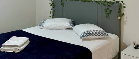 A neatly made bed with white linens and a dark blue blanket, a white bedside table with a remote control, and a grey rug on the floor.