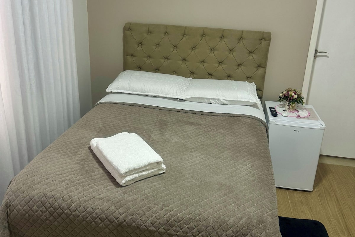 A neatly made bed with a quilted bedspread, a folded white towel, and a small bedside table with a remote control and a vase of flowers.