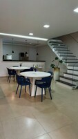 Una cocina moderna con una isla central, un comedor con mesas redondas y sillas azules, y una escalera con barandillas de cristal.