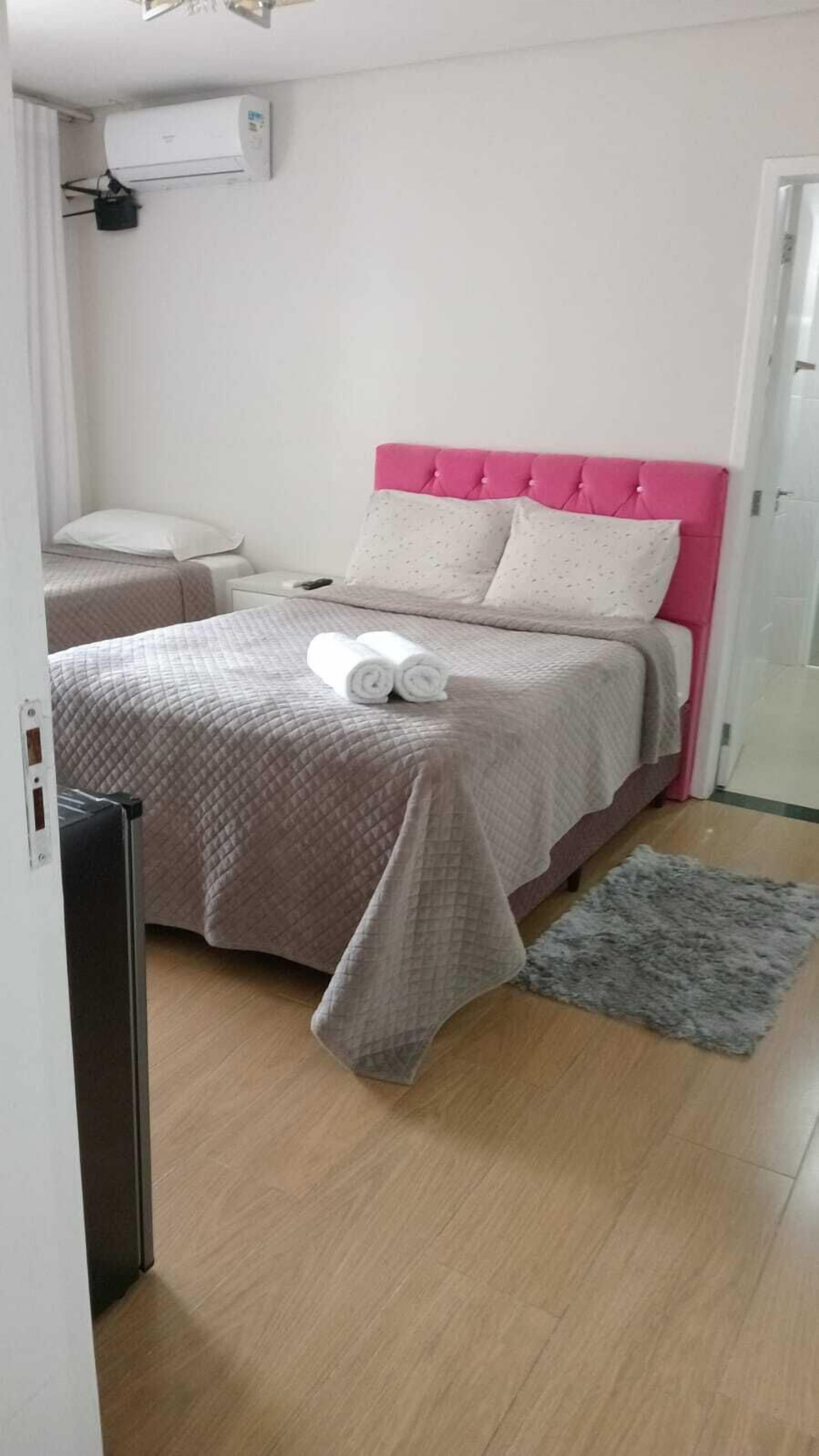 A bedroom with a pink headboard, a bed with a quilted bedspread, two pillows, and a rolled towel on the bed. There is a gray rug on the floor and a white air conditioning unit on the wall.