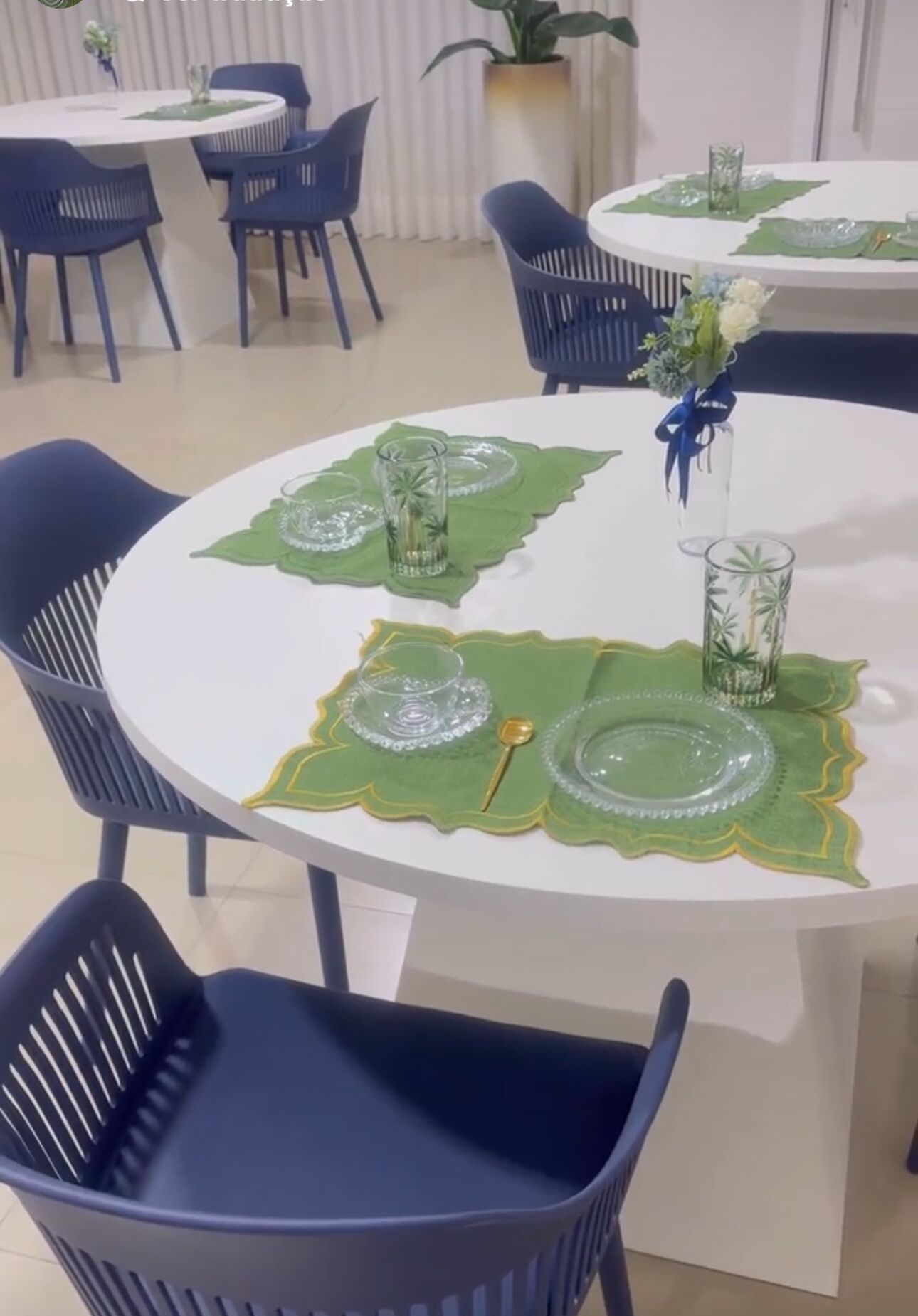 Una mesa redonda de comedor con individuales verdes, cristalería transparente y un jarrón con flores.