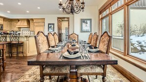 A dining room with a large wooden table set for six, surrounded by upholstered chairs with intricate patterns. A chandelier hangs from the ceiling, and the room features large windows with a view of a snowy landscape.