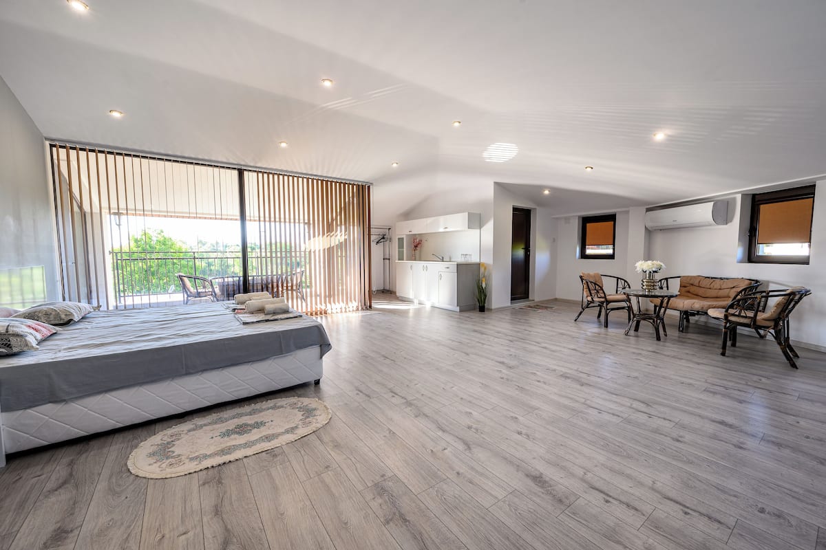 A spacious living area with a large bed, a dining table with chairs, and a kitchen in the background.
