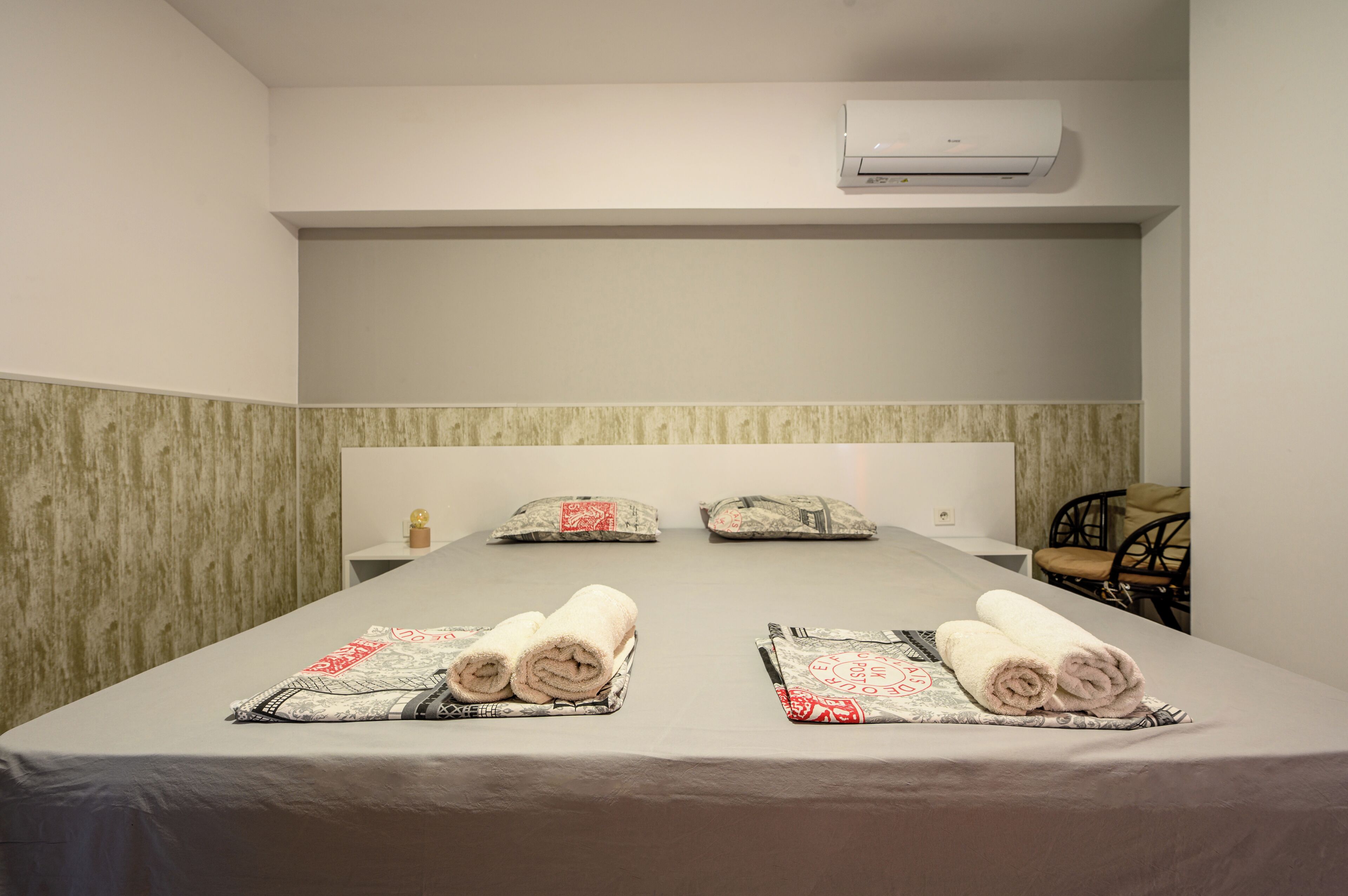 Habitación de hotel con una cama, dos almohadas, una silla, aire acondicionado y una pequeña mesa con una botella.