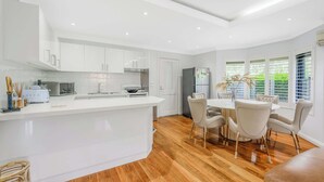 A modern kitchen with white cabinetry, a central island, and a dining area with a round table and chairs. - Maroubra Gem Close to Beach Sleep 16 (Maroubra)