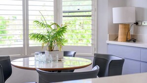 A modern dining area with a round glass table, black chairs, a potted plant, and a white lamp on a shelf. - Maroubra Gem Close to Beach Sleep 16 (Maroubra)